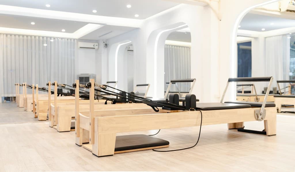 Ahşap reformer pilates aletlerinin sıralandığı, beyaz duvarlı ve geniş perdeli aydınlık bir stüdyo