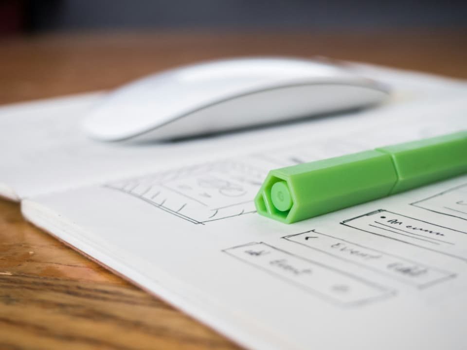 Bir defterdeki el çizimi wireframe taslağının üzerinde duran yeşil bir kalem ve bulanık bir fare